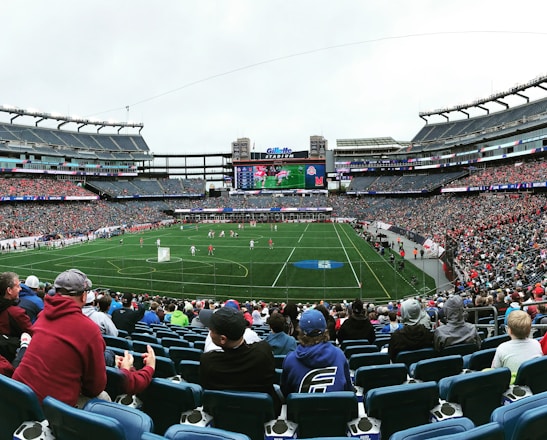 A large sports stadium filled with spectators. The field is green with white markings, and players in red and white uniforms are actively engaged in a game. The stands are packed with people wearing various colored clothing, and some sections of the crowd are more densely populated. Digital screens and advertisements are visible around the stadium, adding to the vibrant atmosphere.