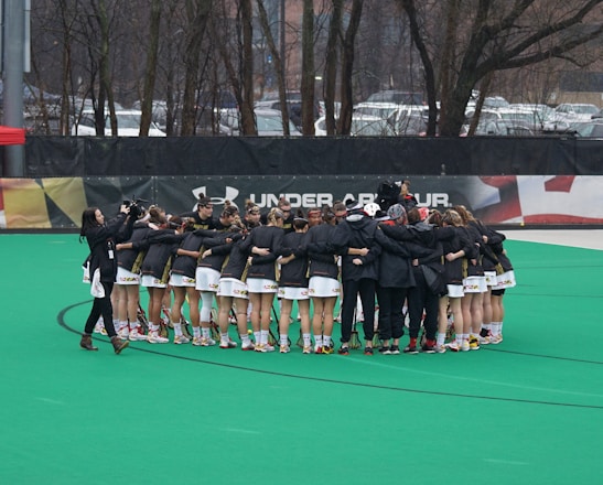 A group of athletes is gathered in a huddle on a sports field, wearing matching uniforms and gear. The ground is green, and there is signage with a brand logo in the background. Trees and parked cars can be seen behind a fence surrounding the field. A person on the left side is capturing the moment with a camera.
