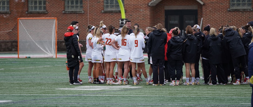 A group of athletes wearing sports uniforms huddles on a grassy field, likely preparing or strategizing for a game. The players are surrounded by additional team members or coaches wearing black jackets. A goalpost and net are visible in the background, along with a brick building.
