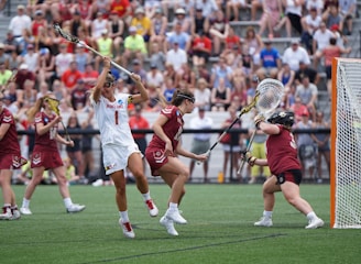 Athletes engaged in a competitive lacrosse match on a grassy field, with one player in white attempting to score against a goalie in burgundy gear. The scene is lively, with a full audience in the background, dressed casually and attentively watching the game.