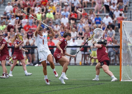 Athletes engaged in a competitive lacrosse match on a grassy field, with one player in white attempting to score against a goalie in burgundy gear. The scene is lively, with a full audience in the background, dressed casually and attentively watching the game.