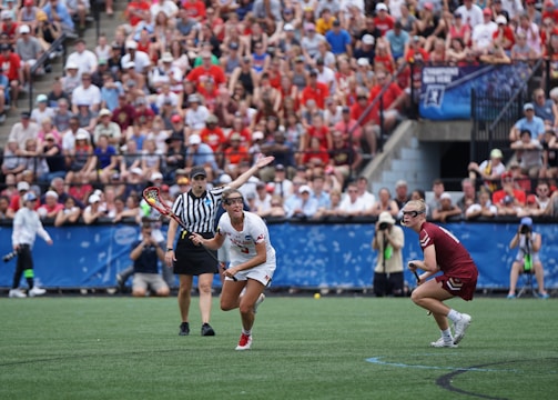A sports event with two athletes engaged in a lacrosse game. One athlete in a white jersey is holding a lacrosse stick, appearing to prepare for a play. A referee in a striped uniform is nearby. The background features a large audience seated in a stadium, with many spectators wearing bright and casual clothing. Some photographers are also visible near the field.