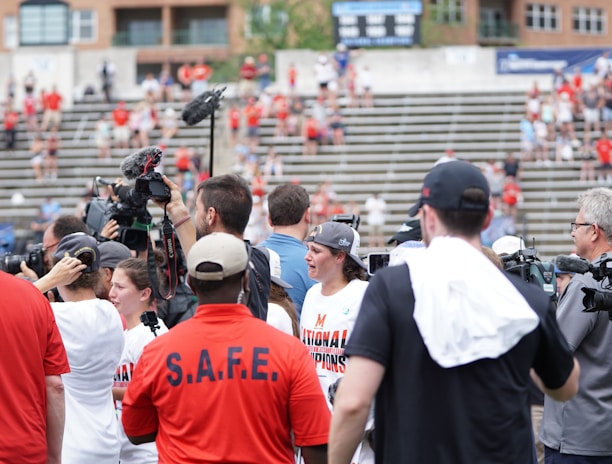A lively gathering in an outdoor sports venue with a group of people, some wearing matching shirts labeled 'National Champions.' Several individuals are holding cameras and microphones, indicating a media presence. The background shows blurred spectators sitting on bleachers.