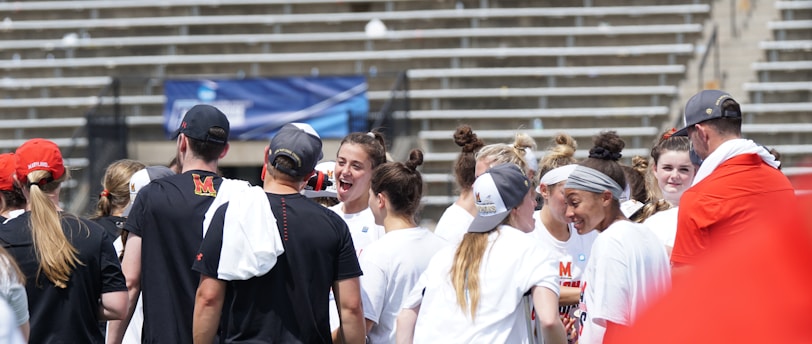 A group of athletes in white shirts with black and red accents are gathered together, engaged in conversation and smiling. They appear to be on a sports field with empty bleachers in the background. The setting is outdoors, and it seems sunny. Some individuals are wearing caps or headbands.