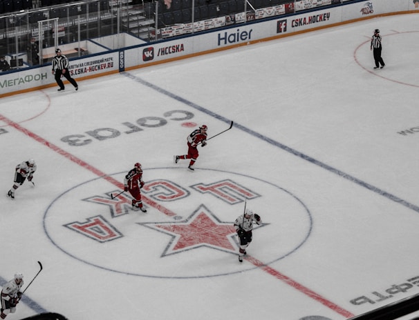 An ice hockey game is underway in an indoor arena with players actively engaged in the sport. Spectators are seated in the stands, while some areas appear sparse. The rink features a large logo at center ice, and electronic advertisements surround the perimeter.