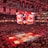 An indoor sports arena filled with spectators and a basketball court illuminated by red lighting. The scoreboards and video screens display promotional content, with numerous banners and championship flags hanging from the ceiling. Players and officials are gathered on the court, possibly in preparation for a game or event.
