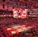 An indoor sports arena filled with spectators and a basketball court illuminated by red lighting. The scoreboards and video screens display promotional content, with numerous banners and championship flags hanging from the ceiling. Players and officials are gathered on the court, possibly in preparation for a game or event.