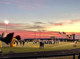 A sports field with a football game unfolding under a vibrant sunset sky, featuring a large inflatable mascot and a scoreboard displaying the score. Players and coaches are visible on the green field, surrounded by a track and some flags waving in the breeze.