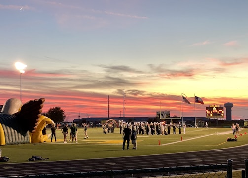 A sports field with a football game unfolding under a vibrant sunset sky, featuring a large inflatable mascot and a scoreboard displaying the score. Players and coaches are visible on the green field, surrounded by a track and some flags waving in the breeze.