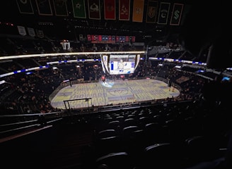 An indoor ice hockey arena with an illuminated scoreboard and a partially filled audience. The ice rink is visible with graphics projected onto the surface, and banners hang from the ceiling. Players are on the ice, and spectators are scattered throughout the stands.