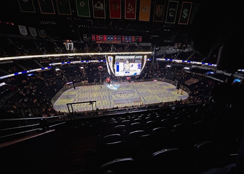 An indoor ice hockey arena with an illuminated scoreboard and a partially filled audience. The ice rink is visible with graphics projected onto the surface, and banners hang from the ceiling. Players are on the ice, and spectators are scattered throughout the stands.