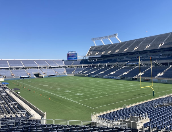 A large, empty sports stadium with blue seats surrounding a well-maintained green grass field. Bright sunlight creates clear shadows, and there are two goal posts on opposite ends of the field. A big electronic scoreboard displays some graphics, and the sky is clear and blue.